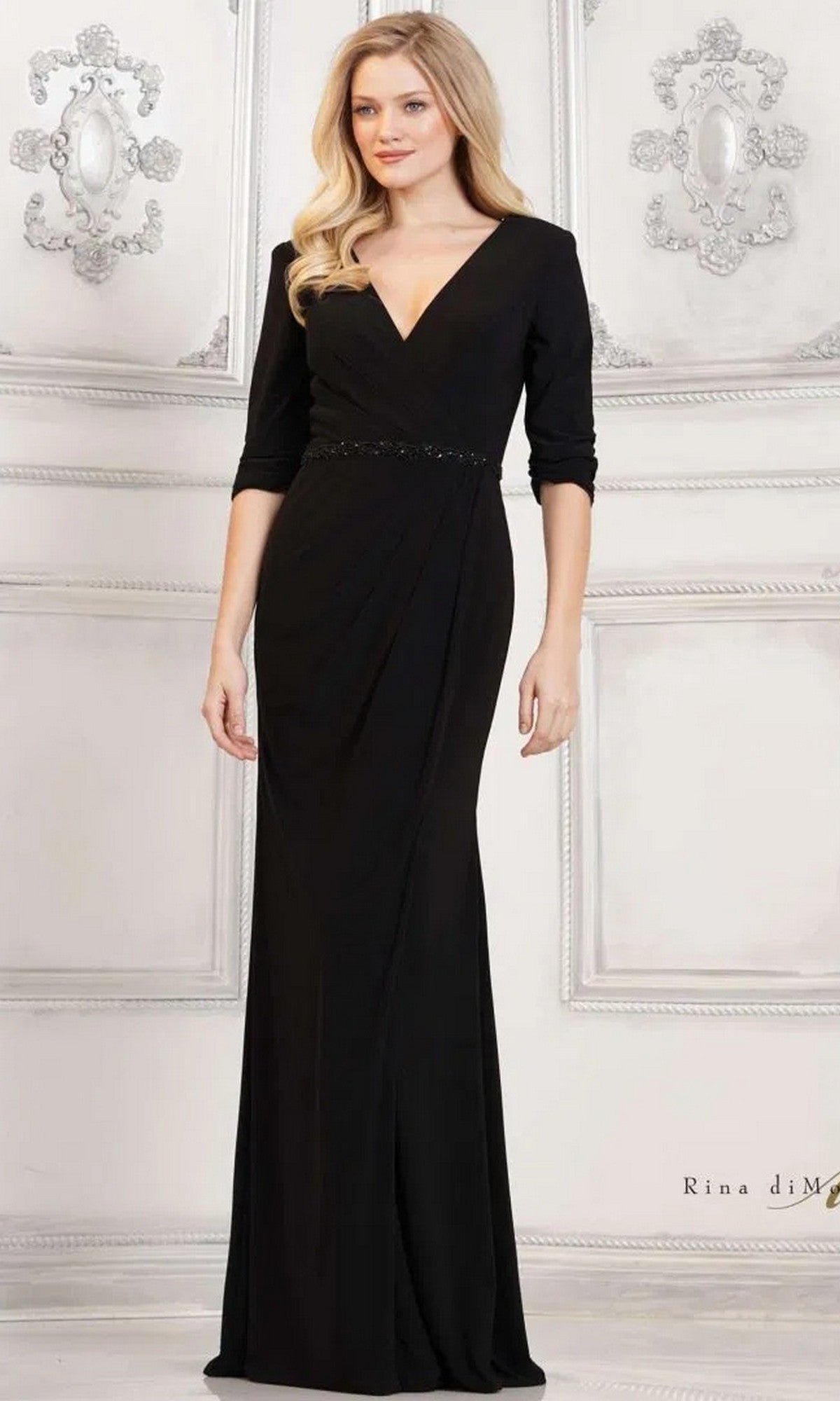 A woman stands against a white ornate wall wearing the black Long Formal Dress: Rina Di Montella RD3109, featuring a V-neck, three-quarter sleeves, and a flattering sheath silhouette.